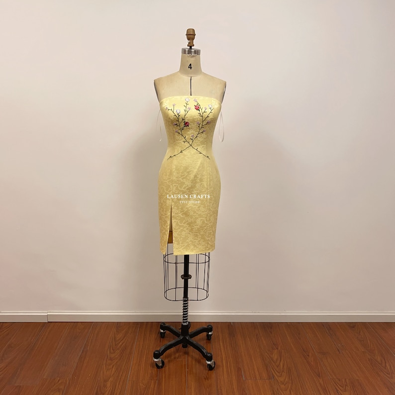 May include: A yellow strapless dress with a floral embroidery design. The dress has a slit on the side and is displayed on a mannequin.
