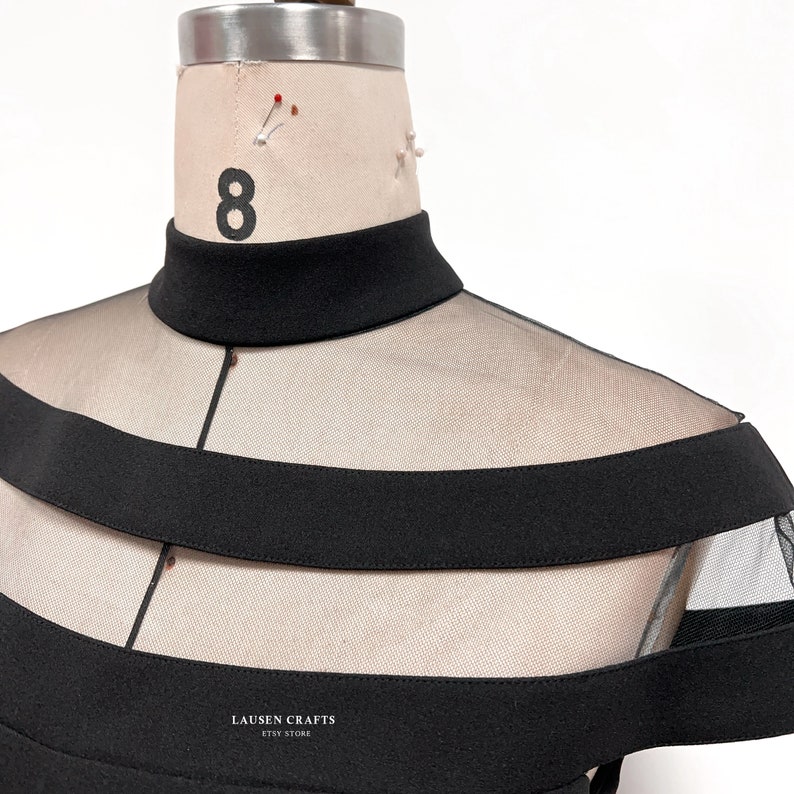 Pu&ograve; includere: Top nero a collo alto con sovrapposizione di rete trasparente e bordatura nera. Il top ha un collo alto e una silhouette aderente. Il testo "LAUSEN CRAFTS ETSY STORE" &egrave; stampato sul davanti del top.