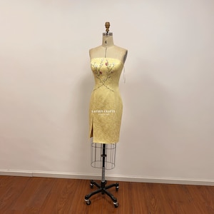 May include: A yellow strapless dress with a floral embroidery design. The dress has a fitted bodice and a straight skirt with a slit on the left side. The dress is displayed on a mannequin.