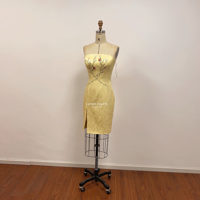May include: A yellow strapless dress with a floral embroidery design. The dress has a fitted bodice and a straight skirt with a slit on the left side. The dress is displayed on a mannequin.