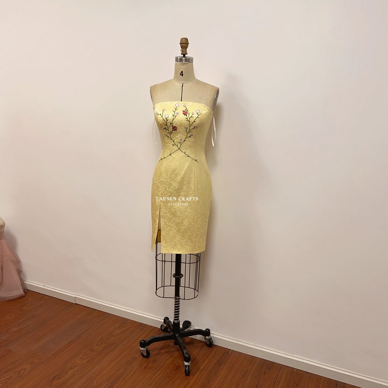 May include: A yellow strapless dress with a floral embroidery design. The dress has a fitted silhouette and a slit on the side. The dress is on a mannequin.