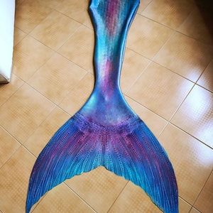 Pode incluir: Uma cauda de sereia em tons de azul, roxo e verde-azulado, com um efeito gradiente. A cauda apresenta um design de barbatana detalhado e está colocada sobre uma superfície ladrilhada. As cores da cauda transitam de um roxo profundo para um azul vibrante.