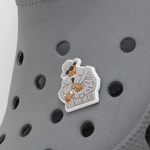 May include: A gray shoe with a cartoon character charm. The charm is of a man wearing a hat, sunglasses, and a coat. The man is holding his hands in front of him.