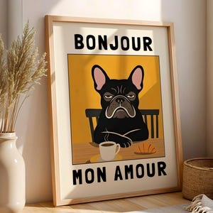 May include: Framed art print featuring a black French Bulldog with a grumpy expression, seated at a table with a coffee cup and croissant. The words "BONJOUR MON AMOUR" are displayed in bold, black letters on a yellow background.