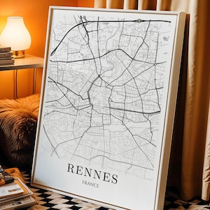 May include: A framed map of Rennes, France, in black and white. The city's streets and roads are depicted in detail. The city name and country are printed in a simple, modern font at the bottom of the map. The frame is white.
