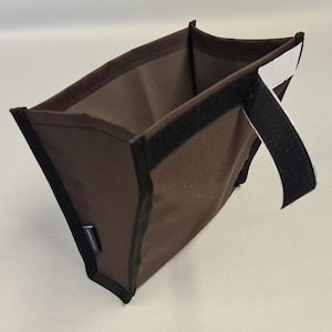Pode incluir: Uma bolsa de tecido marrom com um z&iacute;per preto e uma al&ccedil;a preta. A bolsa tem uma etiqueta branca com texto preto.