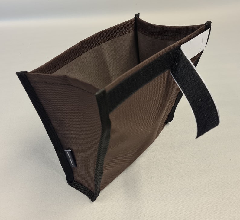 Pode incluir: Uma bolsa de tecido marrom com um z&iacute;per preto e uma al&ccedil;a preta. A bolsa tem uma etiqueta branca com texto preto.