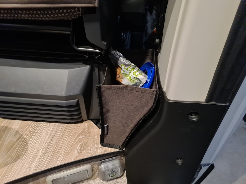 Pode incluir: Um bolso de armazenamento de tecido castanho com guarni&ccedil;&atilde;o preta est&aacute; preso ao lado de um ve&iacute;culo. O bolso est&aacute; cheio de um saco de guloseimas para c&atilde;es e um brinquedo de pl&aacute;stico azul.
