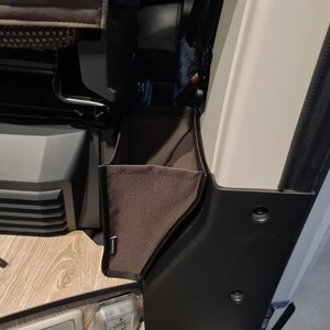 Pode incluir: Um bolso de armazenamento de tecido castanho com um fecho de correr preto e uma guarni&ccedil;&atilde;o preta. O bolso est&aacute; preso ao lado de uma parede branca.