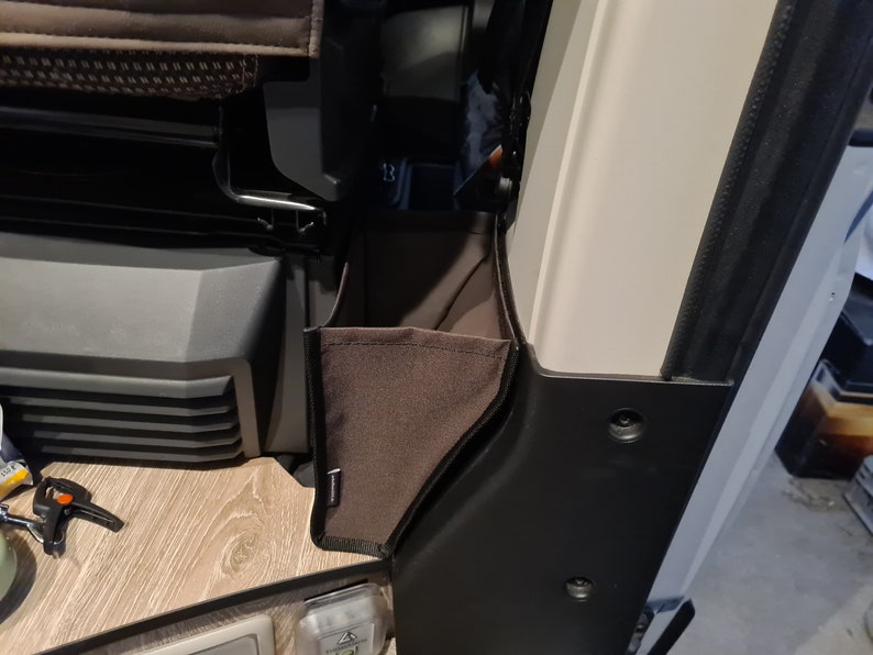 Pode incluir: Um bolso de armazenamento de tecido castanho com um fecho de correr preto e uma guarni&ccedil;&atilde;o preta. O bolso est&aacute; preso ao lado de uma parede branca.