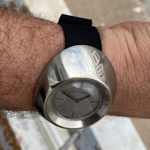 May include: A vintage Pierre Cardin wristwatch with a silver-toned, rounded case and a gray watch face. The watch has a black fabric band. The watch face features thin blue lines at the 12 o'clock position.