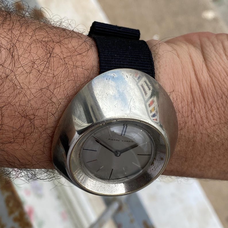 May include: A vintage Pierre Cardin wristwatch with a silver-toned, rounded case and a gray watch face. The watch has a black fabric band. The watch face features thin blue lines at the 12 o'clock position.
