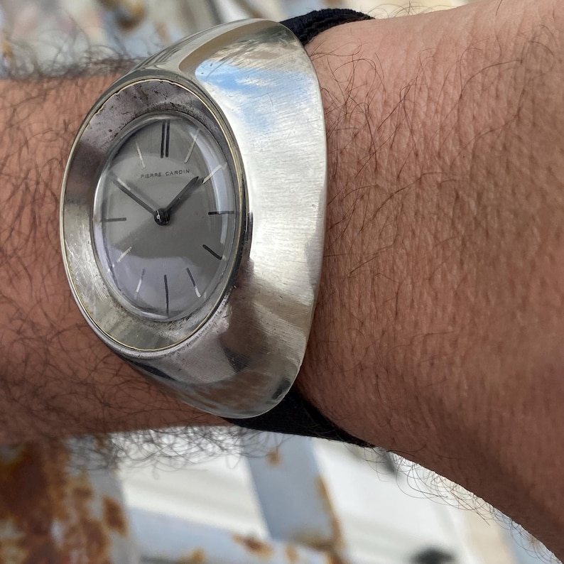 May include: A silver-toned Pierre Cardin wristwatch with a gray face and black hands. The watch has a unique, curved design and is secured with a black band. The watch is a vintage accessory.