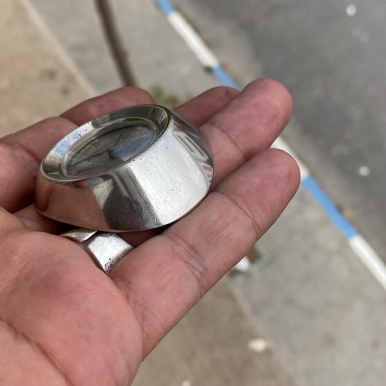 May include: A large, silver-toned ring with a wide band and a flat, oval-shaped top. The ring is held in a hand, showcasing its polished, reflective surface. The ring's design is minimalist and modern, suitable for everyday wear.