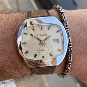 Op de afbeelding: Een vintage Joca Watch met een zilverkleurige kast en een lichtgekleurde wijzerplaat. Het horloge heeft een bruine leren band en een kleine subwijzerplaat. De wijzerplaat toont de tekst "JOCA WATCH" en "17 JEWELS". De datum staat op 13.