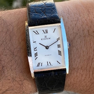 Könnte beinhalten: Eine Vintage-Edox-Armbanduhr mit einem rechteckigen silberfarbenen Gehäuse und einem schwarzen Lederarmband. Das weiße Zifferblatt hat schwarze römische Ziffern, schwarze Zeiger und das Wort "EDOX". Die Uhr ist mit "Swiss Made" gekennzeichnet.