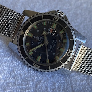 May include: A Mortima Super Datomatic wristwatch with a black dial, silver-tone hands, and a rotating black bezel with white numerals. The watch features a stainless steel mesh strap, a date window at the three o'clock position, and luminous markers.