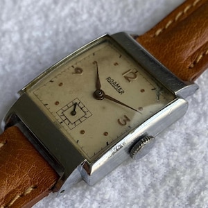May include: Vintage Roamer watch with a rectangular silver case and a light-colored face. The watch features a brown leather band and a small seconds dial. The watch face displays the brand name "ROAMER" and the numbers 3, 6, 9, and 12.