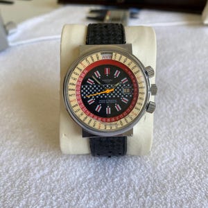 Puede incluir: Un reloj de pulsera vintage con una correa de cuero negro. La esfera del reloj presenta un diseño rojo y negro con texto blanco y un patrón de cuadros. El reloj tiene tres esferas plateadas en el lateral.