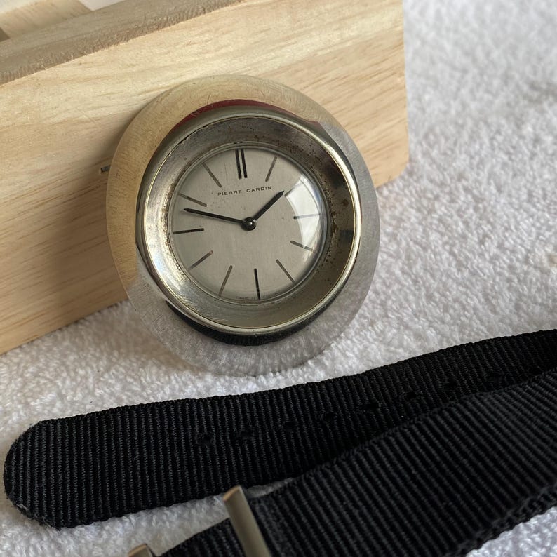 May include: A silver-toned Pierre Cardin wristwatch with a white face and black hands. The watch has a black fabric band. The watch is oval-shaped and sits on a wooden surface.