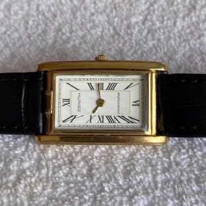 May include: A gold-toned Zenith Cosmopolitan wristwatch with a rectangular face and black leather band. The watch face is white with black Roman numerals and hands. The watch is displayed on a white, textured surface.