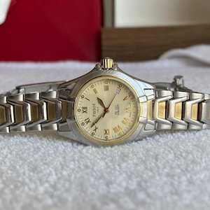 May include: A silver and gold-toned Tissot watch with a cream-colored face. The watch features Roman numeral hour markers and a date display. The band is a combination of silver and gold links. The watch is a PR 100 model.