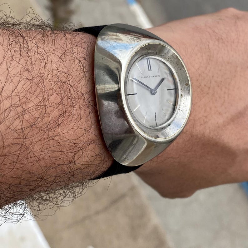 May include: A silver-toned Pierre Cardin wristwatch with a unique oval design. The watch features a light-colored face with silver hands and a black band. The watch is a statement piece.