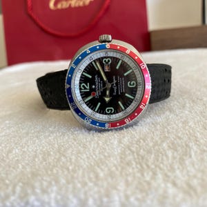 Puede incluir: Reloj Starmaster con esfera negra, bisel giratorio rojo y azul y correa negra. La esfera presenta agujas e indicadores luminosos, con la fecha a las 3. El reloj está sobre una superficie blanca.