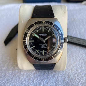 Może przedstawiać: Zegarek na rękę SEAWATCH w stylu vintage z czarną tarczą, srebrną ramką i czarnym gumowym paskiem. Tarcza zegarka wyświetla datę oraz tekst "SEAWATCH 23 JEWELS WATERPROOF 60m 200 TESTED". Zegarek jest prezentowany na białej poduszce.