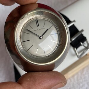 May include: A vintage Pierre Cardin wristwatch with a silver-tone case and a black nylon strap. The watch face is light gray with black hands and markers. The watch is a classic design.