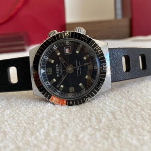 Puede incluir: Un reloj Sicura vintage con esfera negra, agujas luminosas y bisel giratorio. El reloj presenta una correa de cuero negro y está marcado "25 JEWELS" y "SUPERWATERPROOF". La esfera tiene números arábigos y una ventana de fecha.