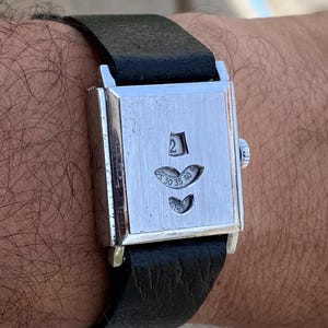 May include: A rectangular silver-tone wristwatch with a black leather strap. The watch face features a brushed metal finish with three distinct cutouts displaying numerical indicators, including the number 2 and a scale ranging from 25 to 40.