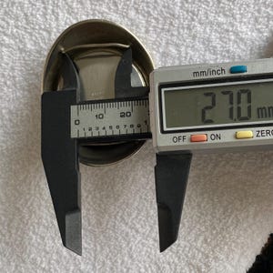 May include: A silver-colored, oval-shaped object is measured with a digital caliper. The caliper displays a measurement of 27.0 mm. The object has a shiny, metallic appearance.