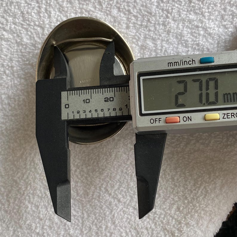 May include: A silver-colored, oval-shaped object is measured with a digital caliper. The caliper displays a measurement of 27.0 mm. The object has a shiny, metallic appearance.