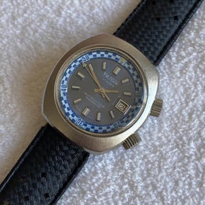 Könnte beinhalten: Eine Vintage-Armbanduhr mit grauem Zifferblatt, blau-weißen Akzenten und einem schwarzen Armband. Das Zifferblatt zeigt den Markennamen "FRUSAN" und den Text "AUTOMATIQUE INCABLOC". Die Uhr hat eine Datumsanzeige bei 3 Uhr.