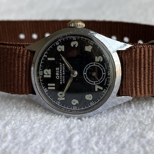 Puede incluir: Un reloj de pulsera Oris vintage con esfera negra, caja plateada y correa de tela marrón. La esfera del reloj muestra números y agujas blancos, con el texto "ORIS WATERPROOF ANTI-SHOCK". Un reloj clásico.