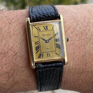 May include: A gold rectangular wristwatch with a black leather band. The watch face is gold with black Roman numerals and the words "Chermidor Paris". The watch band is textured.