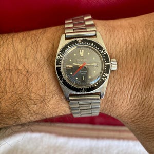 Könnte beinhalten: Eine silberfarbene Vintage-Armbanduhr mit schwarzem Zifferblatt und drehbarer Lünette. Die Uhr verfügt über ein Datumsfenster, Leuchtzeiger und den Markennamen "DUWARD". Das Armband ist aus silberfarbenem Metall.
