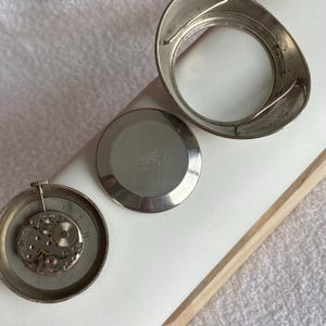 May include: Disassembled vintage watch components, including a silver-toned bezel, back, and movement. The watch parts are arranged on a white surface, showcasing the intricate internal gears and mechanisms of the timepiece.