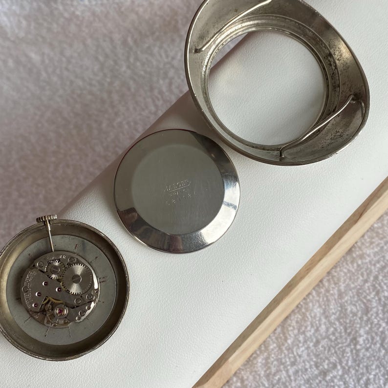 May include: Disassembled vintage watch components, including a silver-toned bezel, back, and movement. The watch parts are arranged on a white surface, showcasing the intricate internal gears and mechanisms of the timepiece.
