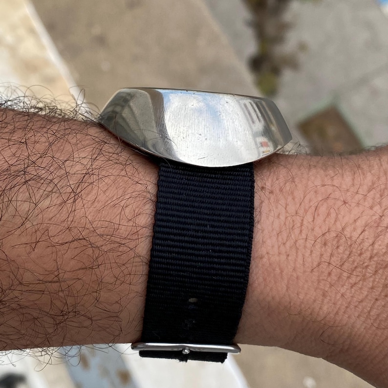 May include: A stainless steel watch with a black nylon strap is shown on a wrist. The watch has a rectangular face with rounded edges. The black strap is secured with a silver buckle. The watch is a classic accessory.