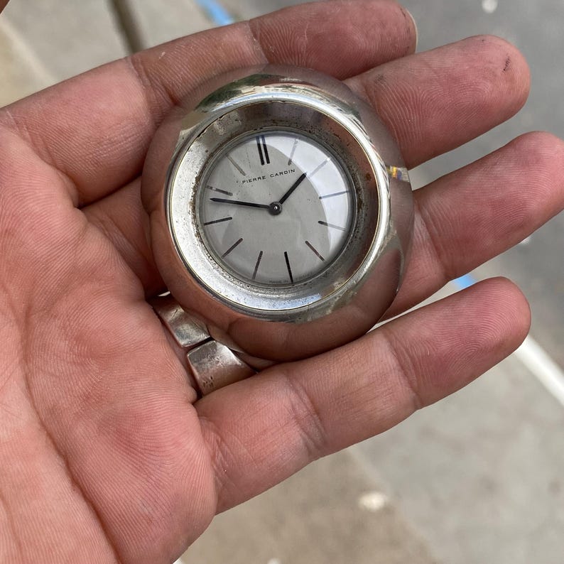 May include: A silver-toned Pierre Cardin wristwatch with a round face and a light gray dial. The watch has black hands and hour markers. The watch is held in a hand.