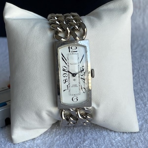 May include: A vintage Thermidor wristwatch with a rectangular white face and silver chain-link band. The watch face features black numerals and hands, with the brand name and "U Swiss Antichoc" printed below the center.