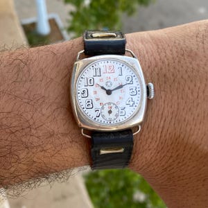 May include: A silver-toned cushion-shaped wristwatch with a white dial, black Arabic numerals, and a red number 12. The watch features a smaller sub-dial for seconds, a black leather strap, and a side crown, displaying a 24-hour inner scale.