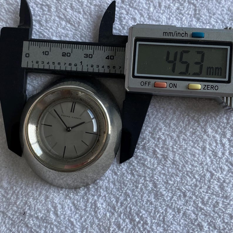 May include: A silver-toned Pierre Cardin watch with a light-colored face, measured at 45.3 mm using a digital caliper. The watch has a minimalist design with silver hands and markers.