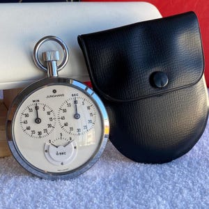 Op de afbeelding: Een zilverkleurige Junghans-stopwatch met een witte wijzerplaat met twee hoofdwijzerplaten voor minuten en seconden, een kleinere 1/10 seconde boogwijzerplaat, een opwindkroon met ring en een bijbehorend zwart, gestructureerd beschermtasje.