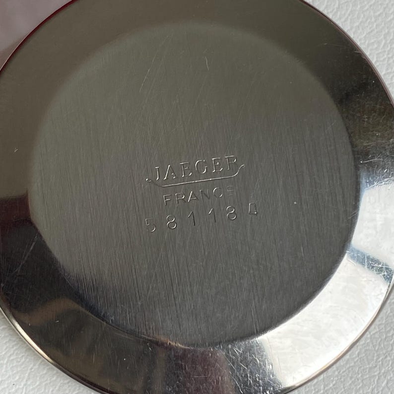 May include: A close-up of a silver-colored, circular object with the text "JAEGER FRANCE 581184" etched into the surface. The object has a brushed metal finish and appears to be a watch component.