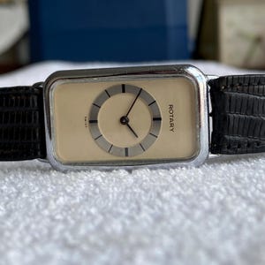 May include: A vintage Rotary watch with a rectangular cream face and black hands. The watch has a silver bezel and a black leather strap. The word "ROTARY" is printed on the face. The watch is a classic design.