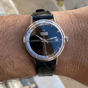 Op de afbeelding: Een vintage polshorloge met een zwarte wijzerplaat en een zilverkleurige lunette. Het horloge heeft een zwarte leren band en de woorden "MICONOS" en "Elegance" zijn zichtbaar op de wijzerplaat. De datum wordt weergegeven als "22".