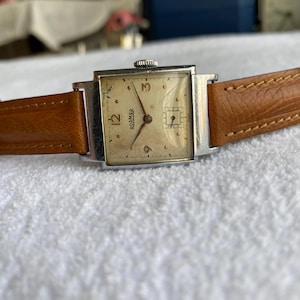 Könnte beinhalten: Vintage Roamer-Uhr mit quadratischem, silberfarbenem Gehäuse und hellem Zifferblatt. Die Uhr hat ein braunes Lederarmband und zeigt die Zeit mit Ziffern und Zeigern an. Auf dem Zifferblatt steht das Wort "ROAMER".